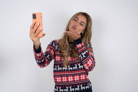 Téléchargez les photos : Femme caucasienne portant chandail de Noël souffle baiser d'air à la caméra du smartphone et prend selfie, envoie mwah via appel en ligne. - en image libre de droit