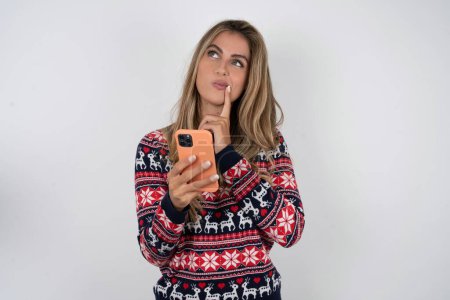 Téléchargez les photos : Femme caucasienne portant un pull de Noël pense profondément à quelque chose, utilise un téléphone mobile moderne, essaie de composer un bon message, garde l'index près des lèvres. - en image libre de droit
