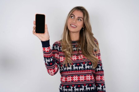 Téléchargez les photos : Femme caucasienne portant chandail de Noël détient un nouveau téléphone mobile et semble mystérieux de côté montre l'affichage vierge de cellulaire moderne - en image libre de droit