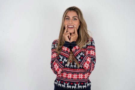 Foto de Mujer caucásica feliz vistiendo suéter de Navidad con sonrisa dentada, mantiene los dedos índice cerca de la boca, dedos señalando y forzando sonrisa alegre - Imagen libre de derechos