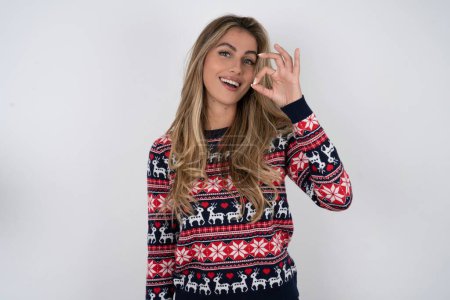 Photo for Caucasian woman wearing christmas sweater hold hand arm okey symbol toothy approve advising novelty news - Royalty Free Image