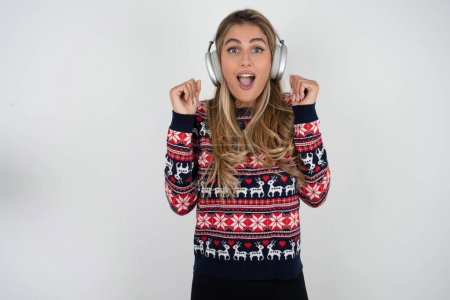 Photo for Emotional caucasian woman wearing christmas sweater exclaims loudly feels like winner raises clenched fists keeps mouth opened wears stereo headphones on ears makes yes gesture, listens favorite music - Royalty Free Image
