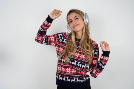 Téléchargez les photos : Femme caucasienne insouciante portant un pull de Noël avec sourire aux dents soulève bras danses mouvements insouciants avec le rythme de la musique écoute de la musique de la playlist via casque - en image libre de droit