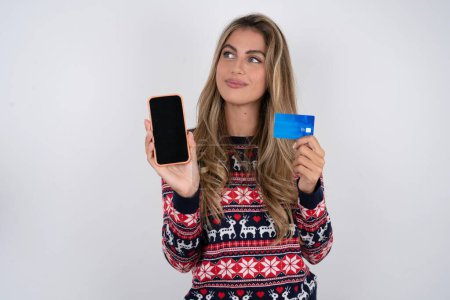 Foto de Mujer caucásica con suéter de navidad celebración tarjeta bancaria dispositivo moderno buscando espacio vacío - Imagen libre de derechos