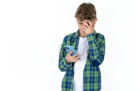 Photo for Handsome caucasian teenager boy on white studio background looking at smart phone feeling sad holding hand on face. - Royalty Free Image