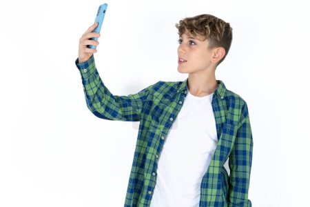Foto de Guapo caucásico adolescente chico en blanco estudio fondo sonriendo y tomando un selfie listo para publicar en su social media. - Imagen libre de derechos
