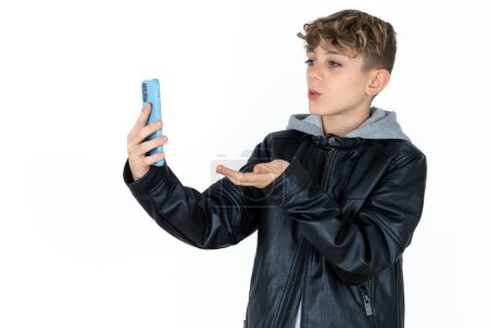 Téléchargez les photos : Beau garçon adolescent caucasien sur fond blanc studio souffle baiser d'air à la caméra du smartphone et prend selfie, envoie mwah via appel en ligne. - en image libre de droit