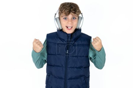 Photo for Emotional handsome caucasian teenager boy on white studio background exclaims loudly feels like winner raises clenched fists keeps mouth opened wears stereo headphones on ears makes yes gesture, listens favorite music - Royalty Free Image