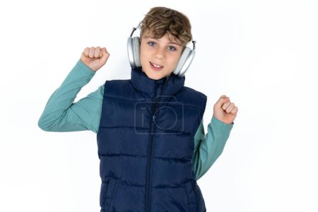 Photo for Carefree handsome caucasian teenager boy on white studio background with toothy smile raises arms dances carefree moves with rhythm of music listens music from playlist via headphones - Royalty Free Image