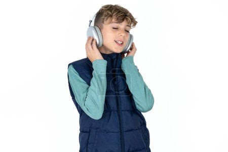 Téléchargez les photos : Beau garçon adolescent caucasien sur fond de studio blanc avec des écouteurs sur la tête, écoute de la musique, profiter de la chanson préférée avec les yeux fermés, tenant la main sur casque. - en image libre de droit