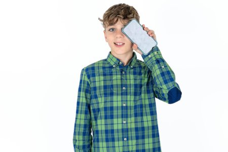 Foto de Guapo caucásico adolescente chico en blanco estudio fondo celebración moderno teléfono inteligente que cubre un ojo mientras sonríe - Imagen libre de derechos