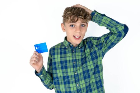 Téléchargez les photos : Impressionné drôle beau garçon adolescent caucasien sur fond blanc studio main sur la tête tenant la carte bancaire - en image libre de droit