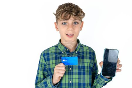 Téléchargez les photos : Photo d'adorable beau garçon adolescent caucasien sur fond de studio blanc tenant la carte de crédit et Smartphone. Réservé aux achats en ligne - en image libre de droit