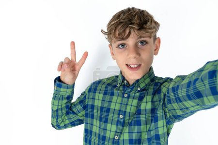 Photo for Positive handsome caucasian teenager boy on white studio background take selfie make v-sign - Royalty Free Image