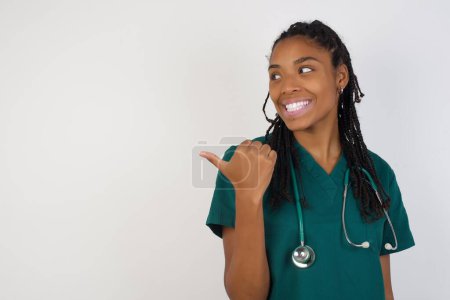 Photo for Omg concept. Stupefied african american doctor woman with surprised expression, opens eyes and mouth widely, points aside with thumb, shows something strange on gray background. Advertisement concept. - Royalty Free Image