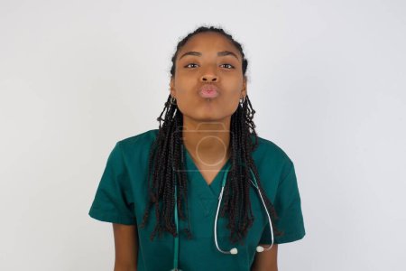Téléchargez les photos : Agréable jeune médecin féminin portant un uniforme médical, garde les lèvres comme aller embrasser quelqu'un, a une expression heureuse, visage grimaçant. Debout dedans. Concept de beauté. - en image libre de droit