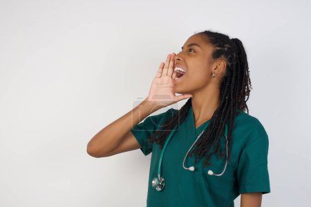 Foto de Africano americano médico mujer de pie contra gris fondo perfil ver, buscando feliz y emocionado, gritando y llamando para copiar espacio. - Imagen libre de derechos
