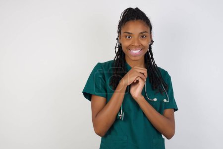 Foto de Dreamy encantadora joven afroamericana médico mujer con expresión agradable, mantiene las manos cruzadas cerca de la cara, emocionado por algo agradable, posa sobre fondo gris. - Imagen libre de derechos