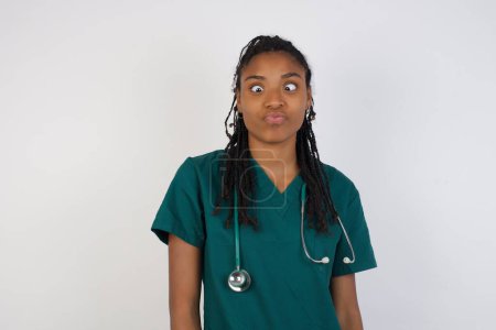 Photo for Funny comic african american doctor woman crosses eyes, with awkward expression has fun alone, puts lips, makes grimace, foolishness after all day. - Royalty Free Image