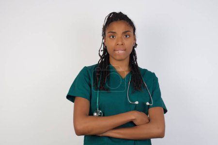 Foto de Retrato aislado de una joven y elegante doctora afroamericana mordiéndose la boca y pareciendo preocupada y asustada cruzando brazos, preocupación y duda. - Imagen libre de derechos