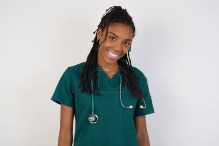 Téléchargez les photos : Belle femme médecin souriant regardant à la caméra portant l'uniforme médical et debout contre le mur gris. - en image libre de droit