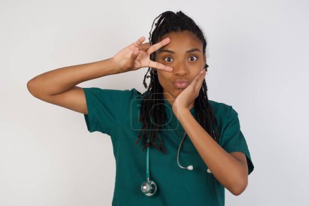 Photo for Leisure lifestyle people person celebrate flirt coquettish concept. Beautiful doctor woman wearing medical uniform showing v-sign near eyes wearing casual clothes standing against gray wall. - Royalty Free Image