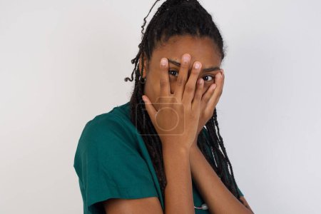 Foto de Hermosa joven cafrican americana médico mujer cubriendo su cara con sus manos y mirando hacia fuera con un ojo entre sus dedos de pie en el interior. Asustado de algo o de alguien. - Imagen libre de derechos