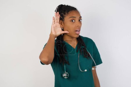 Téléchargez les photos : Oh mon Dieu ! Amusant étonné afro-américain médecin femme ouvrant la bouche largement, levant les sourcils et éclatant les yeux dans la surprise avec la main près de l'oreille en essayant d'écouter les commérages. - en image libre de droit