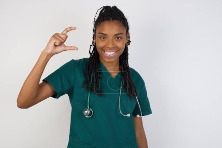 Foto de Médico europeo mujer sobre aislado sobre fondo gris gesto con la mano mostrando tamaño pequeño, símbolo de medida. Sonriendo mirando a la cámara. Concepto de medición. - Imagen libre de derechos