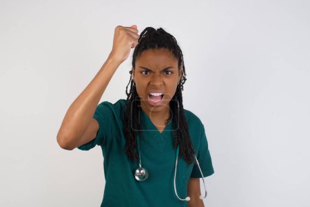Téléchargez les photos : Femme médecin afro-américaine confiante et féroce tenant le poing devant elle comme si elle était prête pour le combat ou le défi, criant et ayant une expression agressive sur le visage. - en image libre de droit