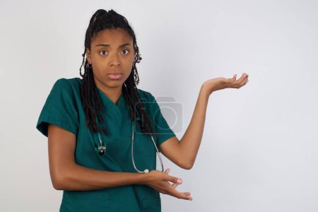 Photo for Beautiful african american doctor girl pointing aside with both hands showing something strange and saying: I don't know what is this. Standing against gray background. Advertisement concept. - Royalty Free Image