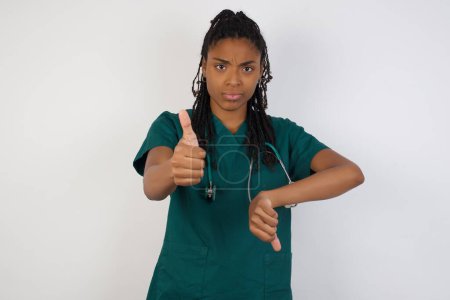 Photo for Pretty young pensive doctor woman making good-bad sign. Displeased and unimpressed wearing green medical uniform and standing against gray wall. - Royalty Free Image