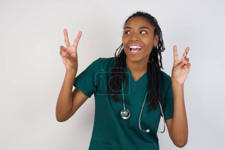 Téléchargez les photos : Photo isolée d'une femme afro-américaine gaie médecin fait signe de paix ou de victoire avec les deux mains, vêtue de vêtements décontractés, se sent cool a sourire aux dents, isolé sur fond gris. Personnes et langage corporel. - en image libre de droit