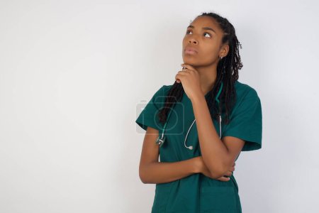 Photo for Portrait of thoughtful smiling african american doctor woman keeps hand under chin, looks sideways, thinking or wondering about something with interest, dressed casually, poses against gray studio. Taking decisions concept. - Royalty Free Image