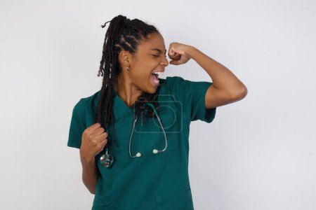 Photo for Attractive young african american doctor woman celebrating a victory punching the air with her fists and a beaming toothy smile over a blue studio background with copy space. - Royalty Free Image