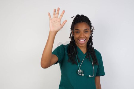 Téléchargez les photos : Portrait d'une jeune médecin afro-américaine joyeuse et chaleureuse qui lève la main et salue, sourit et dit bonjour avec une expression insouciante comme un geste d'adieu ou de bienvenue. - en image libre de droit