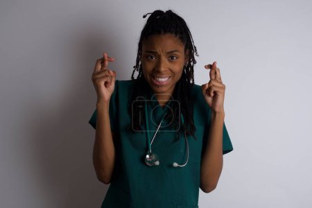 Foto de Hermosa joven afroamericana médico mujer sosteniendo los dedos cruzados con expresión preocupada esperando que su jefe no se dio cuenta de sus errores en el trabajo. - Imagen libre de derechos