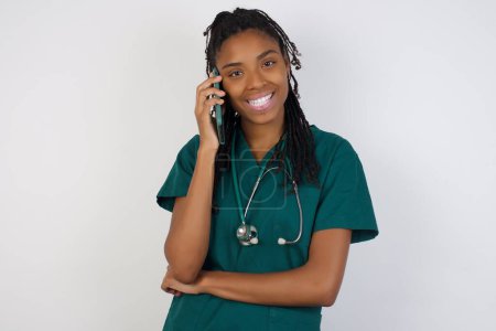 Téléchargez les photos : Portrait d'une jeune femme médecin afro-américaine souriante parlant sur un téléphone portable isolé sur fond blanc. - en image libre de droit