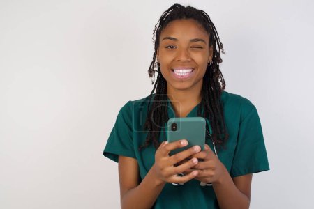 Photo for Pleased african american doctor woman using self phone and looking and winking at the camera over grey background - Royalty Free Image