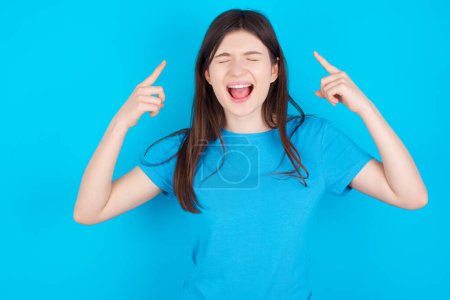 Foto de Foto de una joven caucásica loca usando una camiseta azul aislada sobre fondo azul gritando y señalando con los dedos a los ojos cerrados del pelo - Imagen libre de derechos