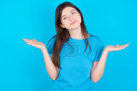 Téléchargez les photos : Jeune fille caucasienne attrayante négligente portant T-shirt bleu isolé sur fond bleu haussant les épaules, oups. - en image libre de droit