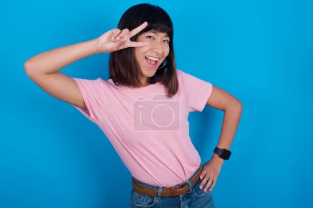 Téléchargez les photos : Jeune asiatique femme portant t-shirt sur fond bleu faisant v-signe près des yeux. Loisirs, coquettes, célébration et concept de flirt. - en image libre de droit