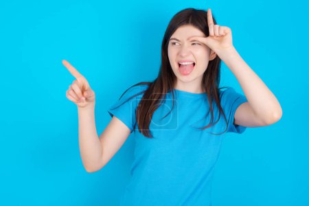 Téléchargez les photos : Jeune fille caucasienne portant bleu T-shirt isolé sur fond bleu montrant signe perdant et pointant vers l'espace vide - en image libre de droit
