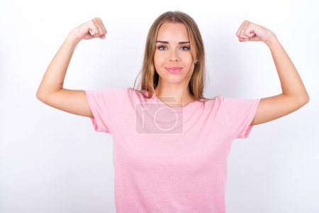 Photo for Waist up shot of Young Caucasian girl wearing pink T-shirt on white background raises arms to show muscles feels confident in victory, looks strong and independent, smiles positively at camera. Sport concept. - Royalty Free Image
