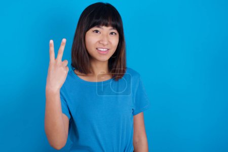 Téléchargez les photos : Jeune femme asiatique portant t-shirt sur fond bleu souriant et regardant amical, montrant numéro deux ou seconde avec la main vers l'avant, compte à rebours - en image libre de droit