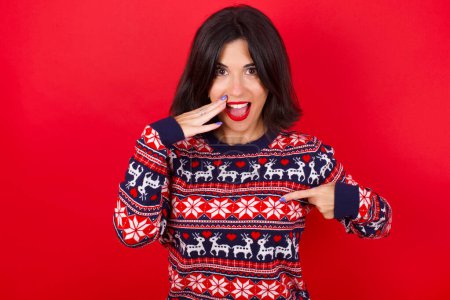 Foto de Sorprendido morena caucásica mujer usando navidad suéter sobre fondo rojo mirada sorprendentemente hacia abajo, indica en blanco con el dedo índice, Modelo asustado - Imagen libre de derechos