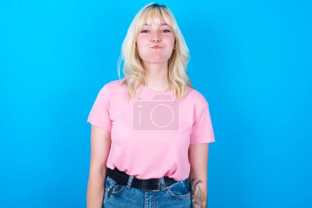 Téléchargez les photos : Fille caucasienne portant t-shirt rose isolé sur fond bleu joues gonflées avec drôle de visage. Bouche gonflée avec de l'air, expression folle. - en image libre de droit