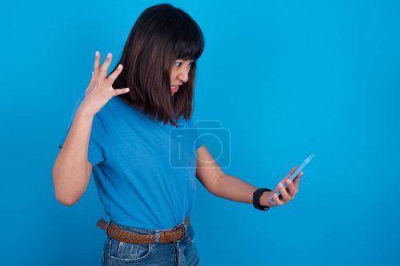 Foto de Foto de indignada joven asiática molesta que usa camiseta contra fondo azul sostiene el teléfono celular, hace llamadas, discute con su colega, expresa emociones negativas. La gente y la ira. - Imagen libre de derechos