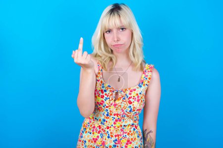 Photo for Caucasian girl wearing floral dress isolated over blue background shows middle finger bad sign asks not to bother. Provocation and rude attitude. - Royalty Free Image