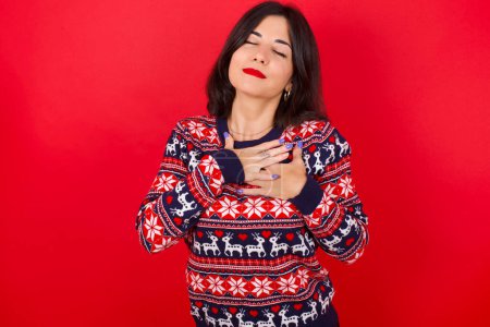 Photo for Brunette caucasian woman wearing christmas sweater over red background smiling with hands on chest with closed eyes and grateful gesture on face. Health concept. - Royalty Free Image